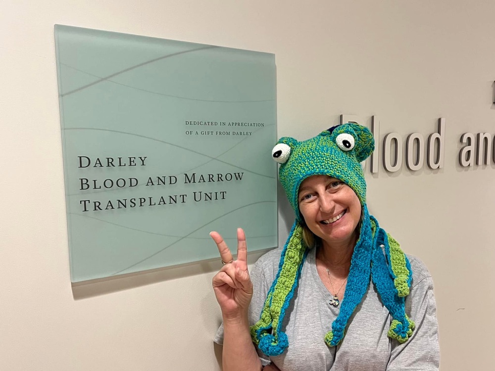 Danell giving a peace sign in a frog hat next to the Blood and Marrow Transplant Unit sign