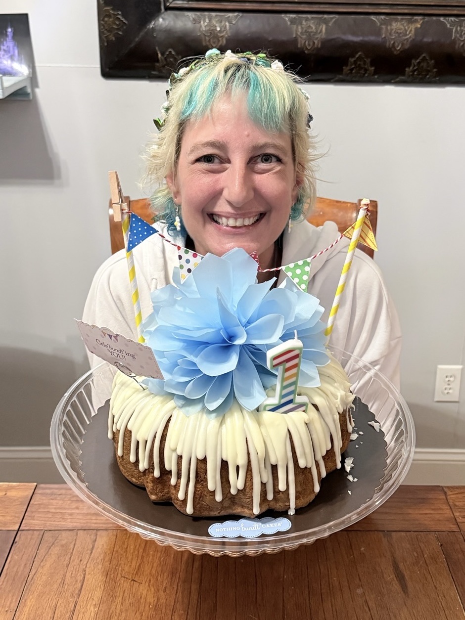 Danell in a flower crown behind a bundt cake with a number one candle, grinning, one year after her transplant